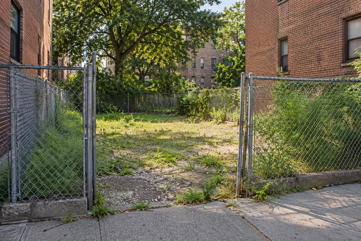 Grassy vacant lot between brick buildings with a fence along the sidewalk