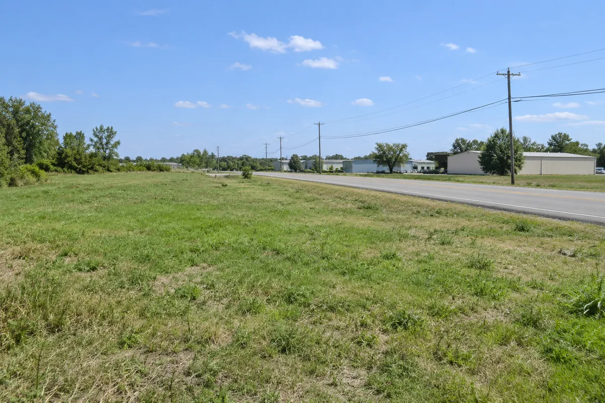 Open grassy site beside a paved road with small industrial buildings in the distance