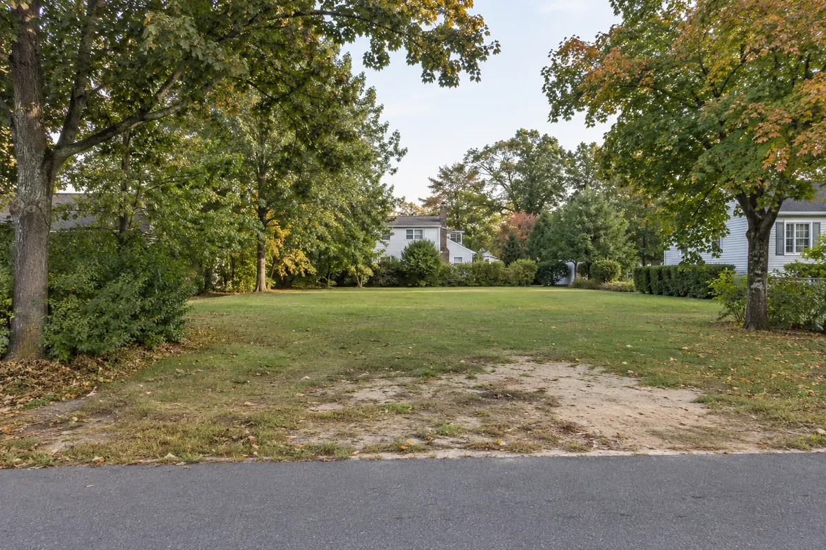 A level grassy vacant lot beside a quiet street with mature trees and a house beyond
