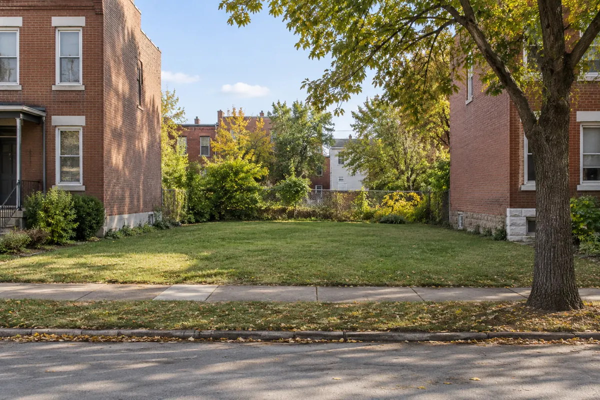 An open grassy city lot between red brick homes, viewed from the sidewalk